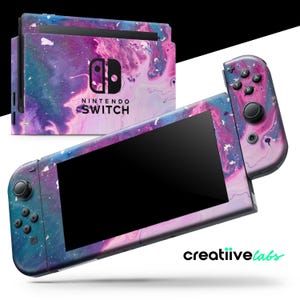 Puede incluir: Una consola Nintendo Switch y sus mandos con un diseño abstracto vibrante en tonos morado, azul y blanco. La consola y los mandos presentan un patrón veteado, similar a una galaxia. El logotipo de Nintendo Switch es visible.