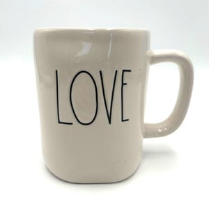 Puede incluir: Taza de cerámica de color crema con asa curva. La palabra "LOVE" está impresa en letras negras estilizadas en la parte delantera de la taza. La taza tiene un diseño simple y minimalista.