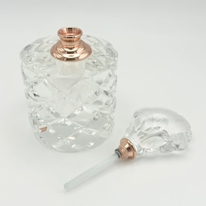 Può includere: Un flacone di profumo in vetro trasparente e sfaccettato con un colletto e un tappo color oro rosa. La bottiglia è cilindrica con un motivo a diamante. Il tappo è staccato, rivelando un applicatore in vetro trasparente.