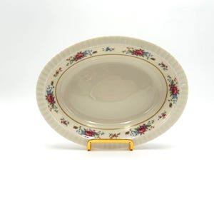 May include: Cream-colored oval serving dish with a floral pattern of red and blue flowers and gold trim. The rim has a ridged texture. The dish is displayed on a gold-colored stand.