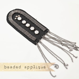 May include: Black beaded applique with silver chain and rhinestone accents. The applique features a curved design with multiple strands of chain hanging from the bottom. The text "beaded applique" is printed on a banner.