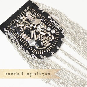 May include: A black fabric applique with silver and black beads and a fringe of silver chains. "beaded applique" is written on a label below the applique.