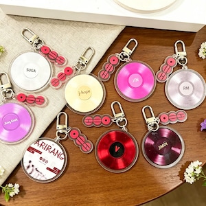 May include: A collection of keychains featuring a vinyl record design in various colors, including pink, red, and silver. Each keychain has a silver clasp and a red accent piece. Some keychains have names like "SUGA" and "Jimin".