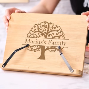 Pode incluir: Um suporte de livro de madeira clara com um desenho de árvore e o texto "Marius's Family". O suporte tem suportes de metal ajustáveis. Uma pilha de livros e uma caneta são visíveis.