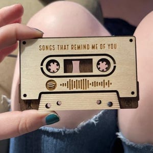 Op de afbeelding: Een houten cassettebandje met de tekst "Songs that remind me of you" en een Spotify-golfontwerp. Het cassettebandje is lichtbruin met een donkerbruin middenstuk en een zwarte rand.