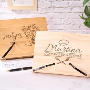 Könnte beinhalten: Zwei Kochbuchständer aus Holz. Einer ist aus Bambus mit "Jocelyn's Family Kitchen" und einem Baumdesign. Der andere ist aus hellem Holz mit "Martina COOKING UP A STORM" und einem Kochhut, einer Gabel und einem Löffel. Beide haben schwarze Metallseitenhalter.