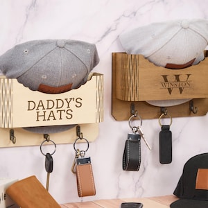 Peut inclure: Deux porte-chapeaux et porte-clés en bois. L'un est étiqueté "DADDY'S HATS" et l'autre porte le nom "Winston". Chaque support a une étagère pour un chapeau et trois crochets pour les clés. Des casquettes de baseball grises et gris clair sont affichées.