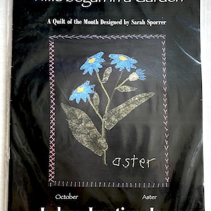 May include: A black and white quilt pattern featuring a blue aster flower with the text "Time began in a Garden" and "A Quilt of the Month Designed by Sarah Sporrer". The pattern is titled "Aster" and is from Indygo Junction, Inc.