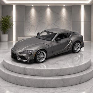 May include: A gray die-cast model of a Toyota Supra sports car. The car is displayed on a marble pedestal with two tiers. The car has silver wheels and a detailed interior. The background is a modern interior with a plant.
