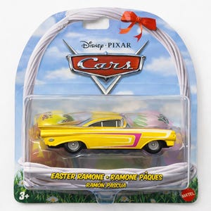 May include: A yellow toy car with pink and purple accents, packaged in a clear plastic case. The car is part of the Disney Pixar Cars series and is labeled "Easter Ramone". The packaging features a blue sky and a red bow.