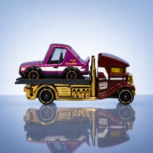 May include: A Hot Wheels toy car hauler with a maroon cab and gold chassis. A purple car with the text "Liberty Walk" is on the hauler. The hauler has the text "Speed Shop" on the side.
