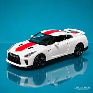 以下が含まれることがあります： 白いダイキャストモデルカー。赤いレーシングストライプがボンネットと屋根の中央にあります。車は黒いホイールとリアスポイラーを備えています。車は反射する青い表面にあります。