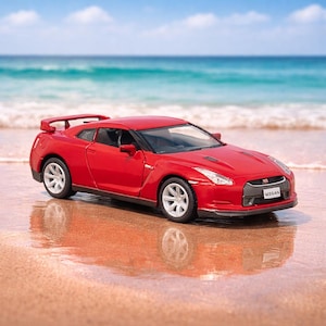 May include: A red Nissan sports car model with silver wheels is parked on a sandy beach. The car has a spoiler and the Nissan logo is visible on the front. The background features the ocean and a blue sky.