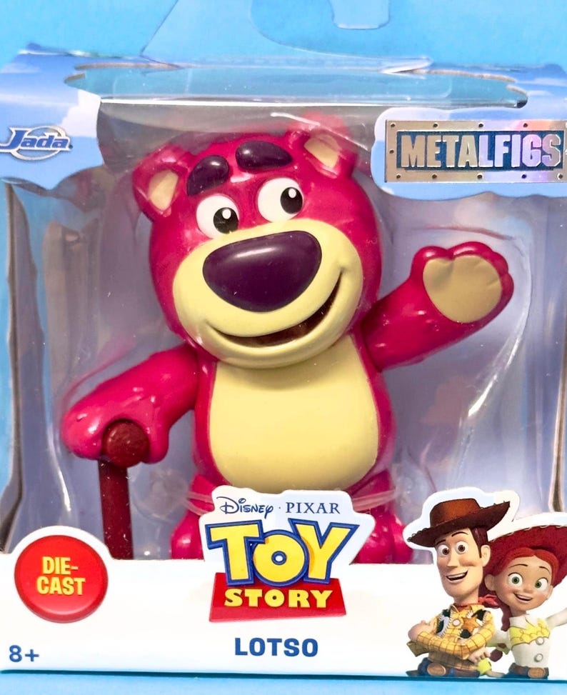 May include: A die-cast Lotso figure from the Disney Pixar Toy Story franchise. The pink bear has a yellow belly and a purple nose. The figure is in a clear plastic package with the Metalfigs logo and the Toy Story logo.
