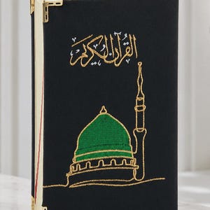 Op de afbeelding: Een zwart hardcover boek met gouden metalen hoekbeschermers en een geborduurd ontwerp op de voorkant. Het borduurwerk toont een gebouw met een groene koepel en een hoge minaret in gouddraad, bekroond met Arabische kalligrafie in goud.