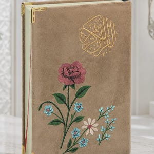 Op de afbeelding: Een met beige fluweel bekleed boek met goudkleurige metalen hoekbeschermers. De voorkant toont goudkleurig geborduurde Arabische kalligrafie boven een bloemmotief bestaande uit een grote roze roos, kleine lichtblauwe bloemen en een witte margriet.