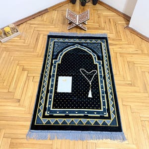 May include: A black prayer rug with a decorative arch design, adorned with gold and blue geometric patterns. A white prayer bead necklace and a small book are placed on the rug. The rug has a fringed border and is set on a wooden floor.