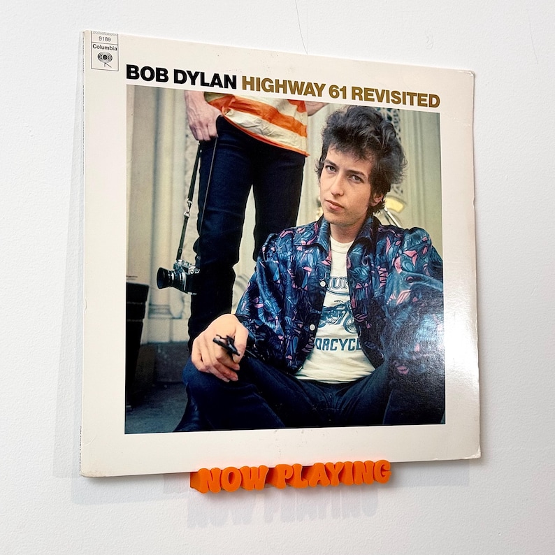 May include: A vinyl record album cover featuring Bob Dylan on the front. The album is titled "Bob Dylan Highway 61 Revisited". The album cover is displayed on a white wall with an orange "NOW PLAYING" sign.