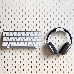 May include: A white wireless keyboard and a pair of black over-ear headphones are mounted on a white pegboard wall.