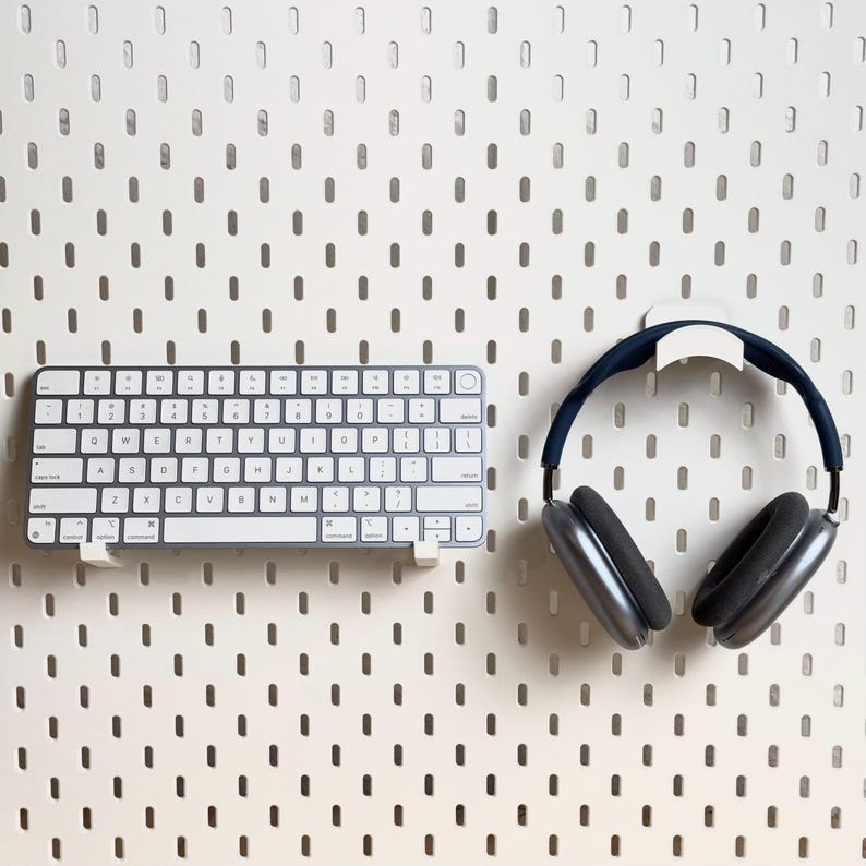 May include: A white wireless keyboard and a pair of black over-ear headphones are mounted on a white pegboard wall.