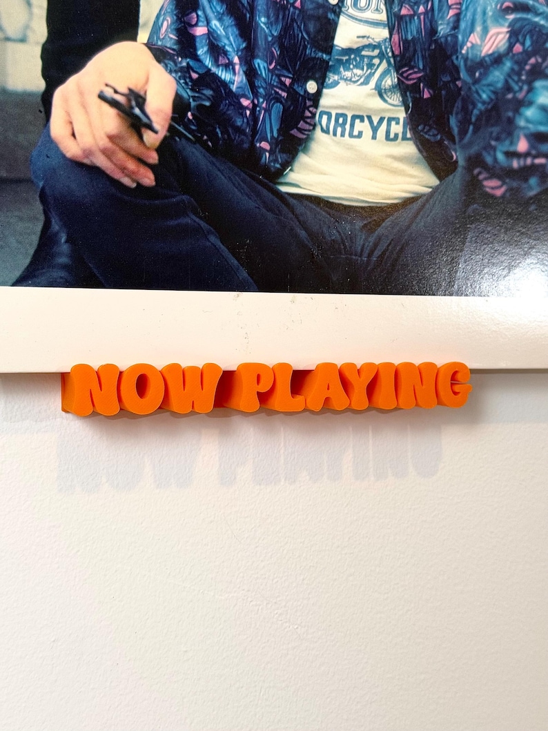 May include: Orange plastic letters spelling "NOW PLAYING" on a white background.