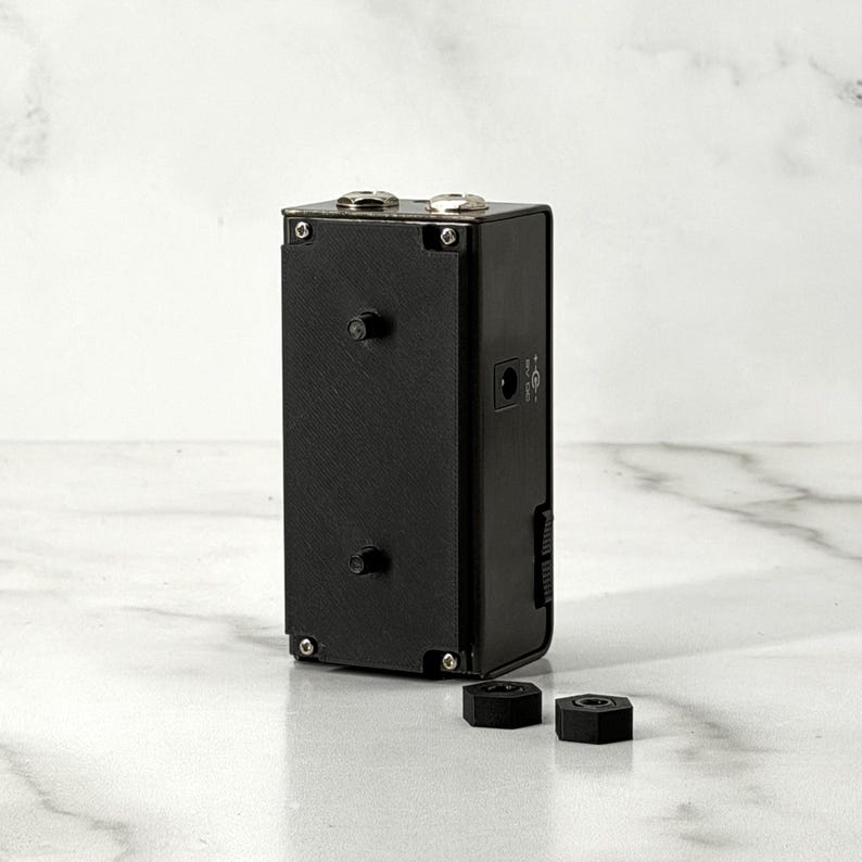 May include: A black rectangular device with two silver-colored knobs on top and several ports on the side. Two black hexagonal nuts are in front of the device. The device is on a white marble surface.
