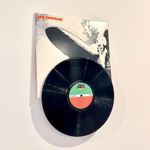 May include: A black vinyl record with a white label that says "Led Zeppelin" and "Atlantic" on it. The record is next to a white album cover with the text "Led Zeppelin" in orange letters. The album cover has a black and white image of an airship.