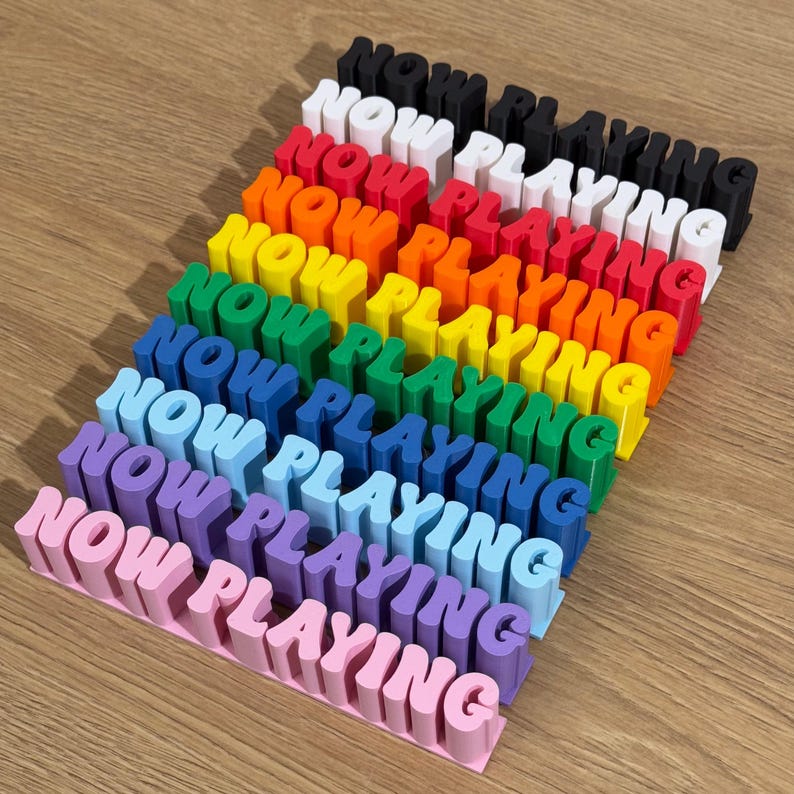 May include: A set of ten "NOW PLAYING" signs in various colors, including black, white, red, orange, yellow, green, blue, light blue, purple, and pink. The signs are stacked on top of each other, with the pink sign at the bottom.