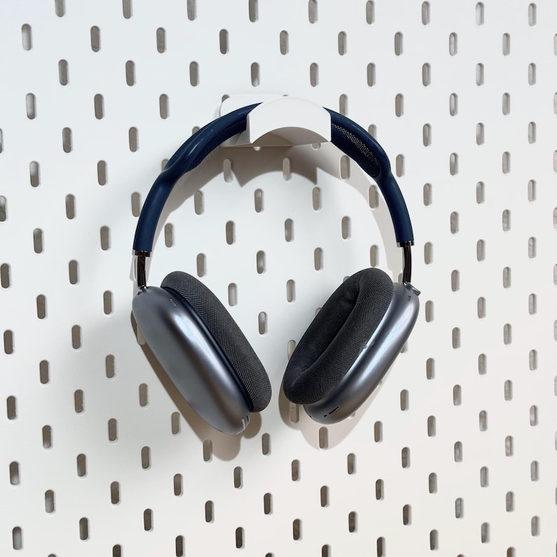 May include: A pair of gray over-ear headphones with a white hook hanging on a white pegboard wall.