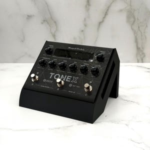 May include: Black ToneX guitar pedal with multiple knobs, buttons, and a digital display. The pedal has the text "ToneX" and "Machine Modeling" printed on it. The pedal is on a black stand.