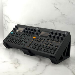 Op de afbeelding: Zwart elektronisch muziekproductieapparaat met talrijke knoppen en een beeldscherm. Het apparaat is voorzien van het label "Digitakt" en is gemonteerd op een zwarte schuine standaard. De standaard is ontworpen om het apparaat te verhogen.