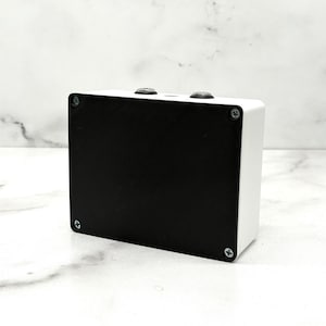 May include: A rectangular electronic enclosure featuring a solid black front panel secured by four silver-colored screws at the corners. The main body of the device is white, and the top edge displays two circular metal components and a small central hole.