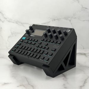 Op de afbeelding: Zwarte elektronische muzieksynthesizer met talrijke knoppen en een klein scherm. Het apparaat is gemarkeerd met "Digitone I" en is gemonteerd op een zwarte standaard. Het ontwerp is strak en modern, geschikt voor muziekproductie.
