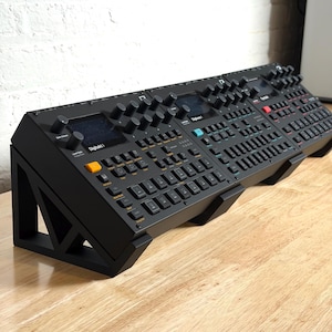 May include: Black electronic music production equipment with multiple knobs, buttons, and screens. The devices are mounted on a black stand. The text "Digitakt" is visible on the front of the device.