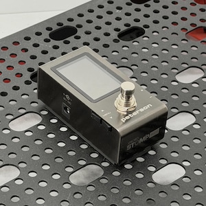 May include: A silver and black Peterson Strobostomp Mini guitar pedal with a screen, a silver button, and the word "Peterson" printed on the front. The pedal is on a black, perforated surface.