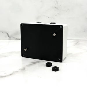 Puede incluir: Una caja rectangular blanca con una base negra y dos perillas negras en la parte superior. La caja está asegurada con tornillos plateados. Dos tuercas hexagonales negras están frente a la caja sobre una superficie de mármol blanco.