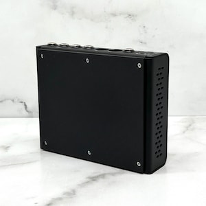 Puede incluir: Un dispositivo electrónico rectangular negro con múltiples puertos y tornillos plateados. El dispositivo tiene una serie de orificios de ventilación en un lado. Es probable que el producto sea un equipo de audio o vídeo.