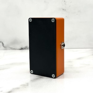 Puede incluir: Dispositivo electrónico rectangular naranja y negro con una entrada jack plateada. El dispositivo tiene cuatro tornillos en la cara negra. Los paneles laterales son naranjas y el panel frontal es negro.