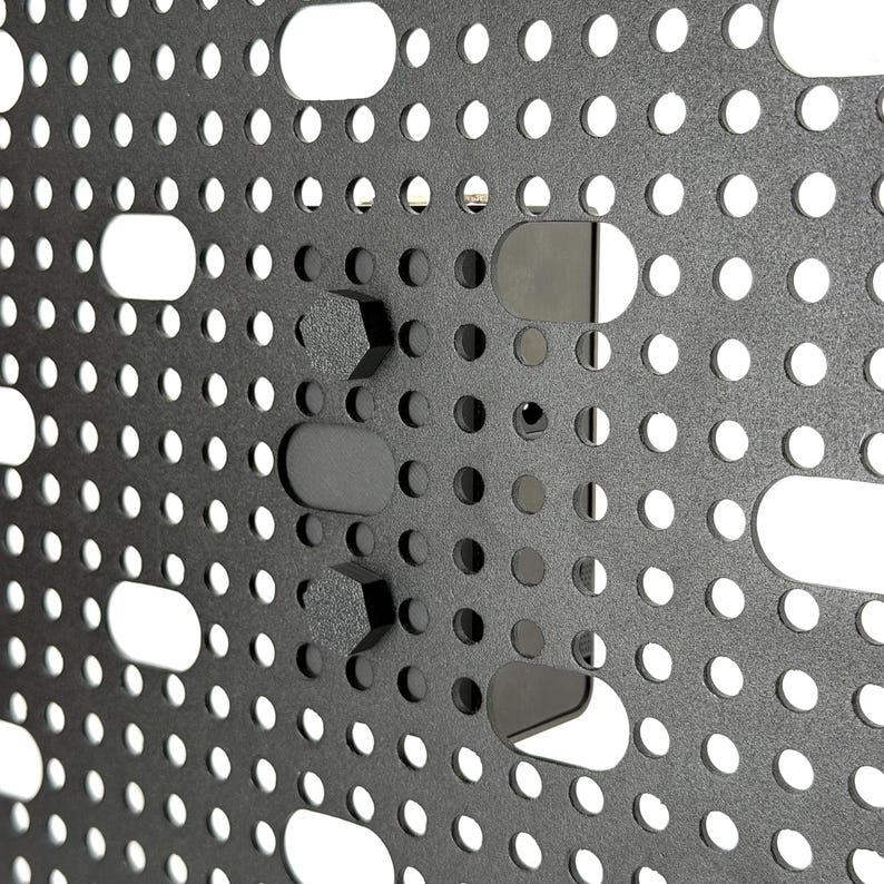 May include: Close-up of a dark gray metal panel with a grid of small, evenly spaced holes and larger, oblong cutouts. Several black, geometric-shaped mounting components are attached to the panel, creating a textured surface.