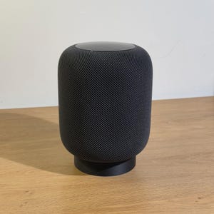 Op de afbeelding: Een zwarte, cilindrische slimme speaker met een gaasrooster en een zwarte basis. De speaker staat op een houten oppervlak.