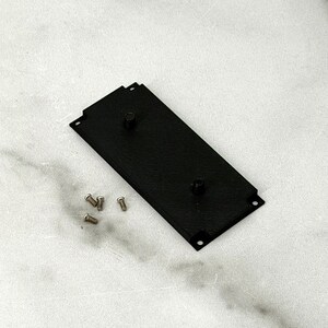 May include: A black rectangular metal plate with two raised cylindrical posts and four small screws. The plate has a textured surface and is positioned diagonally on a light-colored surface.