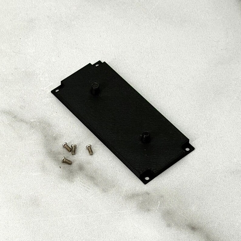 May include: A black rectangular metal plate with two raised cylindrical posts and four small screws. The plate has a textured surface and is positioned diagonally on a light-colored surface.