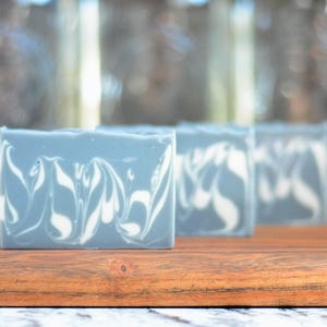 May include: Three rectangular bars of soap featuring a solid slate blue base color with a white marbled swirl pattern throughout the surface. The soap bars are arranged in a row, showcasing a smooth texture and a consistent, clean-cut appearance.