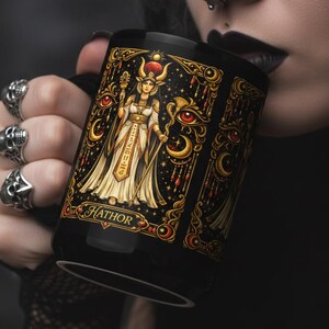 Puede incluir: Taza de cerámica negra con una ilustración detallada de la diosa egipcia Hathor. El diseño incluye detalles dorados, ojos rojos y jeroglíficos. La taza es sostenida por una persona con anillos de calavera.