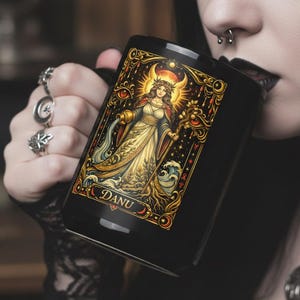 Könnte beinhalten: Schwarze Keramik-Tasse mit einer detaillierten Illustration einer Frau mit goldenem Heiligenschein, Halbmond und dem Wort "DANU". Das Kunstwerk ist mit goldenen und roten Verzierungen umrahmt. Die Tasse wird von einer Person gehalten.