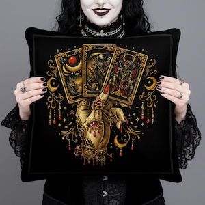 May include: Black square pillow featuring a golden hand holding three tarot cards. The cards depict illustrations of a crescent moon, a grim reaper, and a demon. The hand is adorned with jewelry and a red eye. The pillow has a gothic theme.