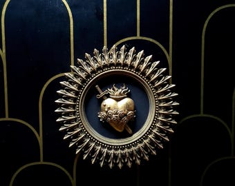 Pode incluir: Uma moldura circular dourada e adornada com um coração dourado em relevo tridimensional, adornado com uma coroa de espinhos e uma adaga. O coração é rodeado por um anel de rosas douradas.