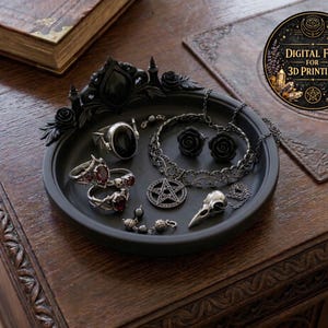 Puede incluir: Una bandeja negra exhibe joyas góticas, incluyendo anillos con piedras rojas, un collar de pentagrama y un colgante de cráneo de pájaro. La bandeja tiene un borde decorativo negro. También se ve un archivo digital para impresión 3D.