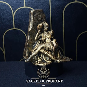 May include: A gold-toned figurine of a woman sitting on a rock throne. She is wearing a cloak and holding a bird. The rock has a Celtic knot design. The base of the figurine is black with a gold circle and eye design. The text "SACRED & PROFANE" is printed on the base.