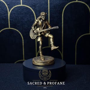 Op de afbeelding: Gouden figuur van een rockmuzikant die gitaar speelt. Het figuur staat op een zwarte basis met een gouden ontwerp. De basis heeft de tekst "SACRED & PROFANE" en "JWL" erop gedrukt.